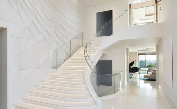 Bright, luxurious modern interior dominated by a sweeping white sculptural staircase with a curved glass balustrade. A textured wave-pattern feature wall rises alongside the stairs, while a double-height circulation space, upper-level landing, recessed dark wall panels and glimpses of a living area create a spacious, gallery-like composition.