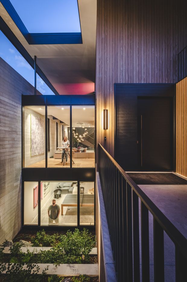 A dramatic contemporary multi-level residential interior/exterior transition viewed from an elevated walkway or balcony. The scene features dark vertical timber wall cladding, black metal-framed glazing, concrete block walls, a covered open-air void, and a recessed dark entry door. Through the glazing, warm-lit living and dining spaces are visible, with a planted courtyard below. The architecture emphasizes strong verticality, layered volumes, and a high-contrast evening atmosphere.