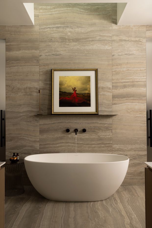A refined bathroom centered on a freestanding white oval bathtub. Large-format beige and grey veined stone tiles cover the walls and floor, with a matching stone shelf, dark wall-mounted tapware, and a framed artwork above the bath. Dark timber or black cabinetry and vertical black handles appear at the sides, while recessed ceiling details provide soft architectural lighting. The overall atmosphere is warm, luxurious, and contemporary.