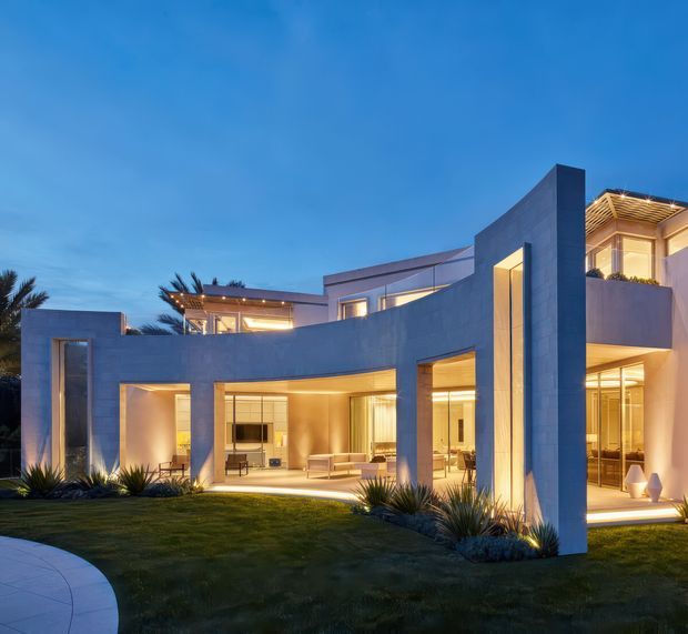 A contemporary multi-level residence photographed at dusk, featuring sculptural curved walls, tall vertical elements, extensive floor-to-ceiling glazing, warm interior illumination, and a landscaped lawn with architectural planting. The facade combines smooth pale rendered surfaces with strong geometric forms and sheltered outdoor living areas.