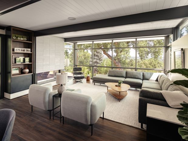 A spacious contemporary living room with a large grey sectional sofa, upholstered lounge chairs, a low timber coffee table, and a neutral area rug. Full-height framed glazing provides views of mature trees, while dark exposed structural beams contrast with a white panelled ceiling. Built-in dark shelving and cabinetry line the left wall, and dark timber flooring runs throughout the room.