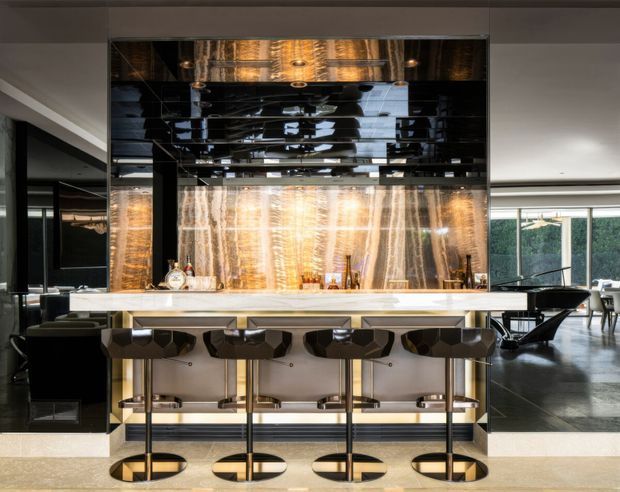 A luxurious contemporary home bar with a long pale stone island and four sculptural dark bar stools. The bar is backed by dramatic veined stone or marble-look panels, reflective black mirror surfaces, integrated shelving, and warm feature lighting. Black cabinetry and glossy finishes create a sophisticated, high-contrast entertaining space, with an adjacent living area visible to the right.