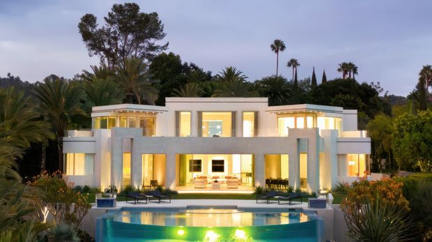 A symmetrical modern luxury residence viewed from the landscaped garden, with a flat-roofed white facade, extensive warm-lit glazing, strong rectangular columns and projecting upper volumes. A swimming pool occupies the foreground, surrounded by tropical and Mediterranean-style planting, with tall palms and cypress-like trees behind the house.
