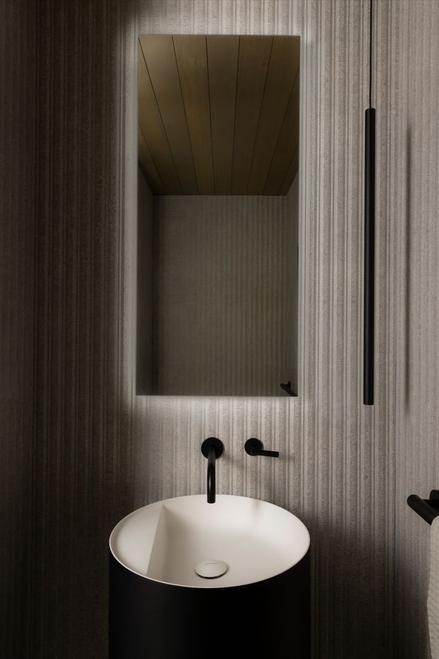A compact, moody bathroom washbasin vignette. A white oval vessel sink sits on a black cylindrical pedestal, paired with matte-black wall-mounted tapware and a tall black vertical handle or accessory. The walls are clad in vertically textured grey patterned material, with a recessed mirrored cabinet above the basin and a warm timber-lined ceiling within the recess. The styling is dark, refined, and minimalist.