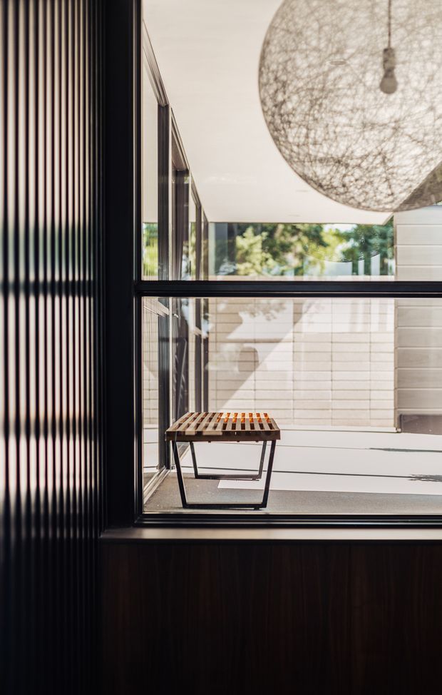 A refined interior view looking through a large black-framed glazed opening toward a bright courtyard or patio. The foreground features dark vertical fluted wall panelling and dark timber joinery, while the courtyard has pale horizontal wall cladding, a simple timber slatted bench, and abundant reflected greenery. A large woven spherical pendant light hangs near the ceiling, adding a sculptural natural texture. The composition emphasizes strong geometric lines, indoor-outdoor connection, daylight, and restrained contemporary detailing.