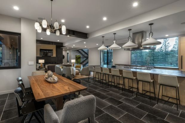 Open-plan contemporary kitchen, dining, and living area with a long stone island and extensive seating beneath multiple pendant lights. The space features dark large-format tiled flooring, dark cabinetry, a timber dining table, grey upholstered chairs, recessed ceiling lights, and large windows overlooking trees. A staircase and living area are visible in the background.