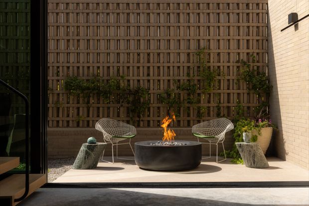 A compact contemporary courtyard terrace viewed from inside through large dark-framed glazing. The paved outdoor seating area features two sculptural wire lounge chairs, irregular stone side tables, and a central round black fire bowl with flames. A tall timber privacy screen with integrated climbing greenery forms the backdrop, while a pale brick wall and potted planting frame the space.