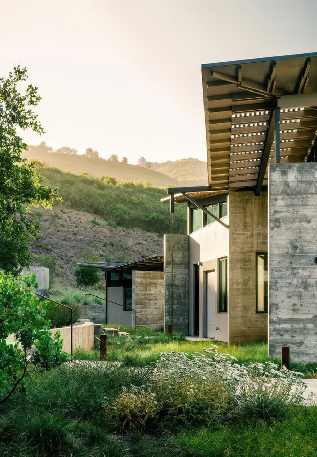Exterior architectural photograph of a modern hillside residence integrated into a landscaped sloped site. The building features exposed concrete walls, dark metal framing, extensive glazing, projecting roof planes, and a prominent open-jointed slatted roof canopy. Native-style planting and terraced paths surround the structure, with rolling hills visible behind it.