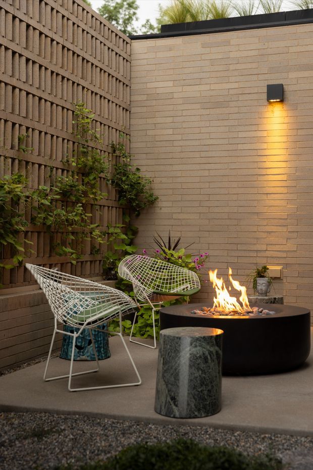 A compact, enclosed outdoor courtyard or patio with a warm contemporary feel. A tan brick wall meets a vertical timber privacy screen covered with climbing greenery. Two white wire-frame lounge chairs surround a black round fire bowl, accompanied by a dark stone cylindrical side table. A wall-mounted light casts a warm glow across the brickwork, while potted plants soften the space.