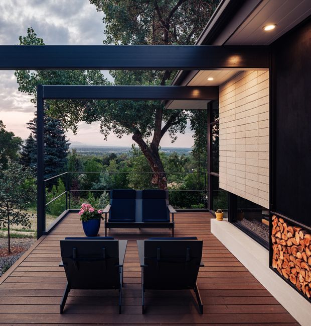 A covered timber deck overlooking a leafy, elevated landscape. The outdoor living area features dark-framed steel or aluminium structural posts and beams, built-in ceiling lighting, a lounge sofa and two armchairs facing a large open fireplace with neatly stacked firewood. Light horizontal wall cladding contrasts with dark wall surfaces and warm timber decking.