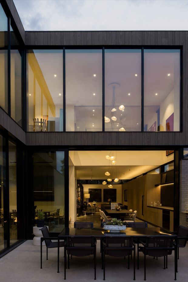 A modern multi-level home viewed from an outdoor dining terrace, with a dark vertical-clad facade, extensive black-framed floor-to-ceiling glazing, and a large open connection between the terrace and the interior dining and kitchen area. Warm recessed lighting and sculptural pendant lights illuminate the dark timber-toned interior, while an upper-level glazed room overlooks the terrace.