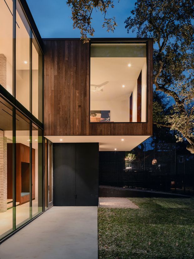 Twilight exterior view of a contemporary multi-level residence with a projecting upper volume clad in dark vertical timber, extensive floor-to-ceiling glazing, slim dark window frames, and a black ground-level entry volume. A covered concrete walkway and landscaped lawn frame the building, while warm interior lighting creates a strong contrast with the blue evening sky.