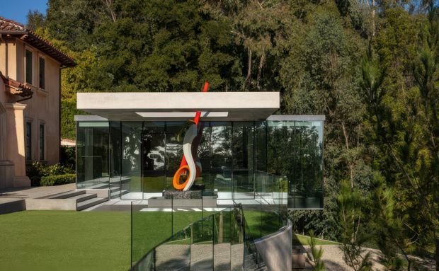 Exterior view of a contemporary glass pavilion or extension set into a landscaped, wooded site. The structure has extensive floor-to-ceiling glazing, a flat projecting concrete roof slab, slender dark frames, and a sculptural red, black, and orange artwork visible at the entrance. A transparent glass balustrade borders a stepped lawn and curved terrace, while part of a more traditional stucco building with a tiled roof is visible at left.