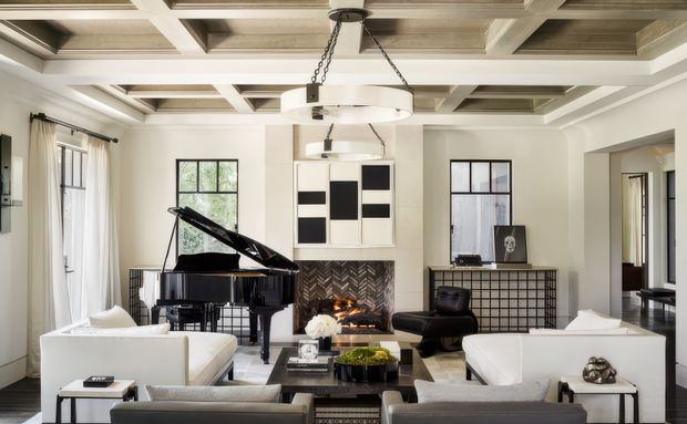 A refined living room with a grand piano, fireplace, lounge seating, and a coffered ceiling. The room features light neutral walls, large black-framed windows, white sofas, a black marble-like coffee table, dark built-in cabinetry, and a suspended circular chandelier. A herringbone-patterned fireplace surround and adjacent black grid-front cabinetry provide visual contrast.