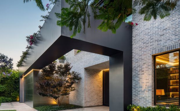 A contemporary residential exterior featuring a dramatic black rectangular portal framing a recessed entry courtyard. The facade combines pale grey brickwork with dark metal cladding, warm concealed architectural lighting, extensive integrated planting, and a large window revealing a warmly lit interior with timber shelving. The image is photographed at dusk and emphasizes strong geometric massing and contrast between the dark structure and light masonry.