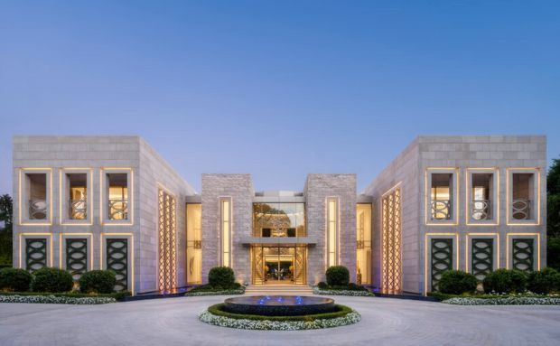 Symmetrical, large-scale contemporary mansion exterior with pale stone-clad wings flanking a recessed glass-fronted central entry. The facade features tall rectangular windows, vertical illuminated recesses, patterned screens, warm interior lighting, manicured hedges, and a circular driveway fountain under a clear blue sky.
