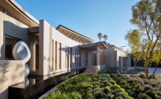 Exterior view of a contemporary low-rise residence with layered horizontal roof forms, projecting eaves, vertical timber-toned cladding, dark framed glazing, and a sculptural translucent feature at the entrance. The building is set within a carefully designed drought-tolerant landscape with shrubs and grasses, under clear daylight.
