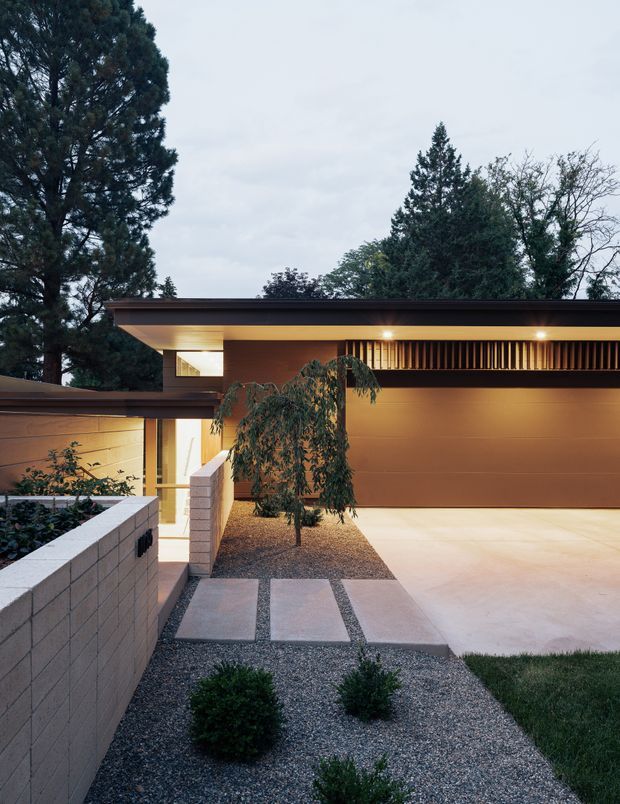 A dusk exterior view of a contemporary single-storey residence, focused on the garage and entry approach. The building has a broad flat roof with deep eaves, warm recessed lighting, smooth brown-toned wall surfaces, a horizontal timber-like louvred ventilation screen, and pale concrete block boundary walls. A gravel garden with clipped shrubs and stepping slabs leads toward the illuminated entry, while mature trees form the background.