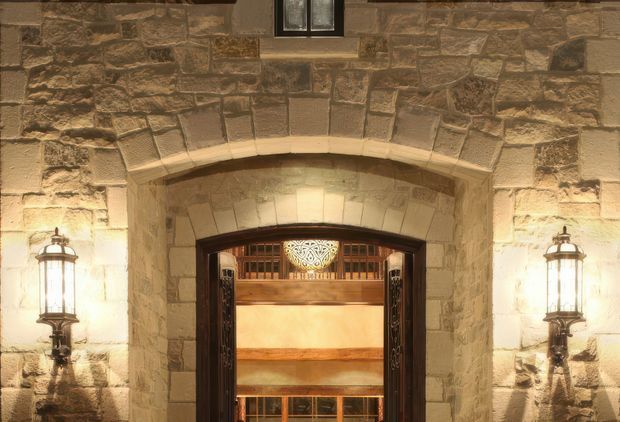 Close-up exterior view of a grand stone building entrance. The facade is constructed from irregular warm-toned masonry with a large arched opening, dark timber-framed doorway, decorative metalwork, and symmetrical traditional wall lanterns. Warm illumination highlights the textured stone and entry surround.
