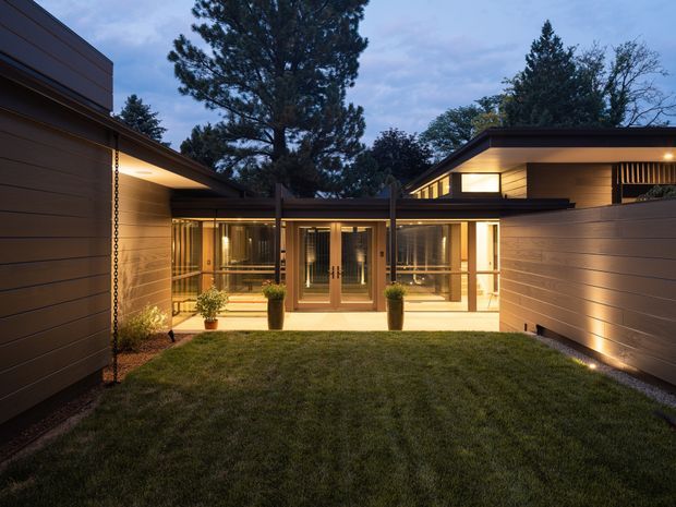 Twilight view of a low-slung contemporary residence arranged around a private grass courtyard. Dark horizontal cladding, broad roof overhangs, extensive warm-lit glazing, and timber-framed doors create a sheltered, inward-facing composition, with mature trees visible beyond.