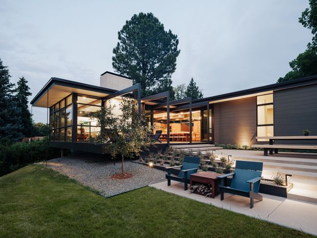 Exterior dusk view of a low-profile contemporary residence with strong horizontal rooflines, extensive floor-to-ceiling glazing, dark cladding, warm interior lighting, and a landscaped outdoor seating area with concrete paving and gravel garden beds.
