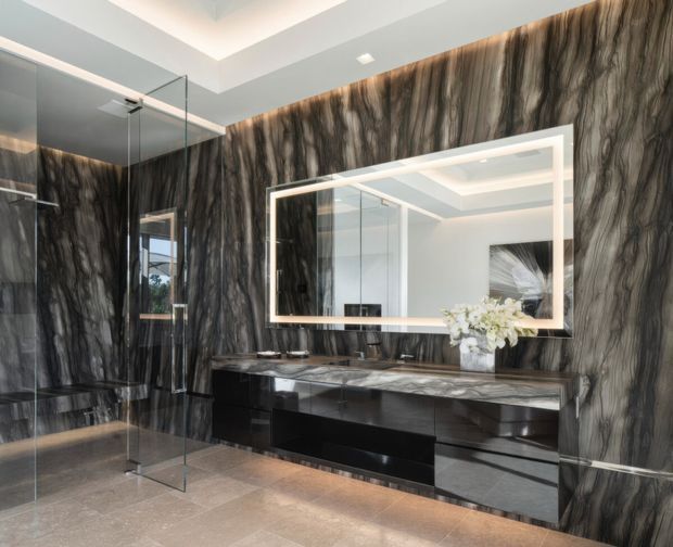 Luxurious contemporary bathroom featuring dramatic dark veined stone-look wall surfaces, a long glossy black floating vanity with integrated basin and drawers, a large illuminated rectangular mirror, frameless glass shower enclosure, and recessed ceiling lighting. The restrained palette of black, charcoal, grey, and white creates a polished, high-contrast finish.