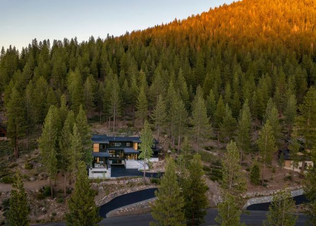 Aerial view of a contemporary multi-level house nestled in a dense coniferous forest on a steep, mountainous site. The dark-clad residence has broad glazing, several roof levels, terraces, and a winding paved driveway with stone retaining walls. The surrounding hillside is illuminated by warm autumn light.