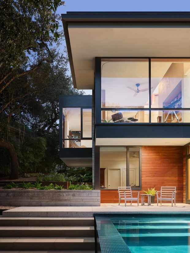 Exterior view of a contemporary multi-level home set among mature trees, showing large glazed openings, dark metal framing, broad roof overhangs, warm timber wall cladding, stepped landscaping, and a swimming pool in the foreground.