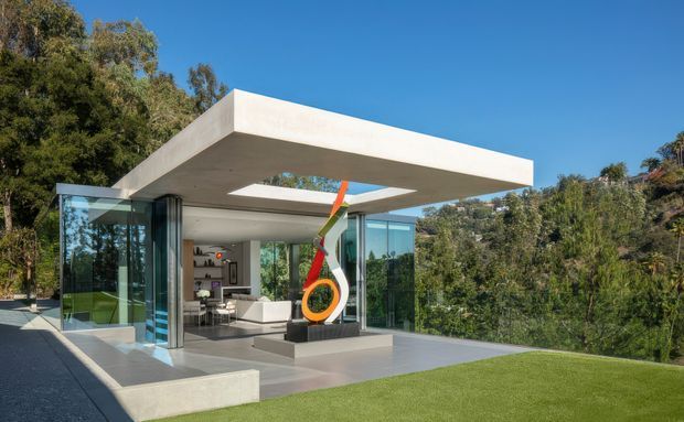 A sleek modern flat-roof pavilion-like house set among mature trees, with extensive floor-to-ceiling glass walls, a large projecting concrete roof slab, and a rectangular roof opening above a covered outdoor terrace. The open-plan interior is visible through the glazing, while a colourful sculptural artwork stands on a dark plinth in the foreground. The scene has crisp contemporary detailing, pale concrete surfaces, and a manicured lawn.