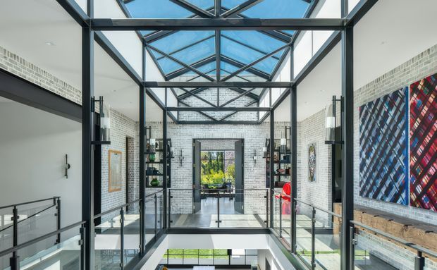 A bright, multi-level interior atrium or landing with a dramatic glazed roof, exposed black steel framing, white painted brick walls, glass balustrades, and a view through to a landscaped exterior. Contemporary artwork and shelving line the walls, while a stair or void opens below, creating a strong sense of height and openness.