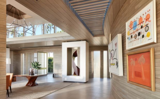 A bright, spacious contemporary living and gallery-like interior with extensive warm timber wall and ceiling panelling, clerestory glazing, and a dramatic slatted ceiling feature. The room includes a sculptural dining table and chairs, a large freestanding display niche with abstract sculpture, artwork on the walls, pale flooring, and indoor planting. The open-plan space has strong natural light and a high, partially vaulted ceiling.
