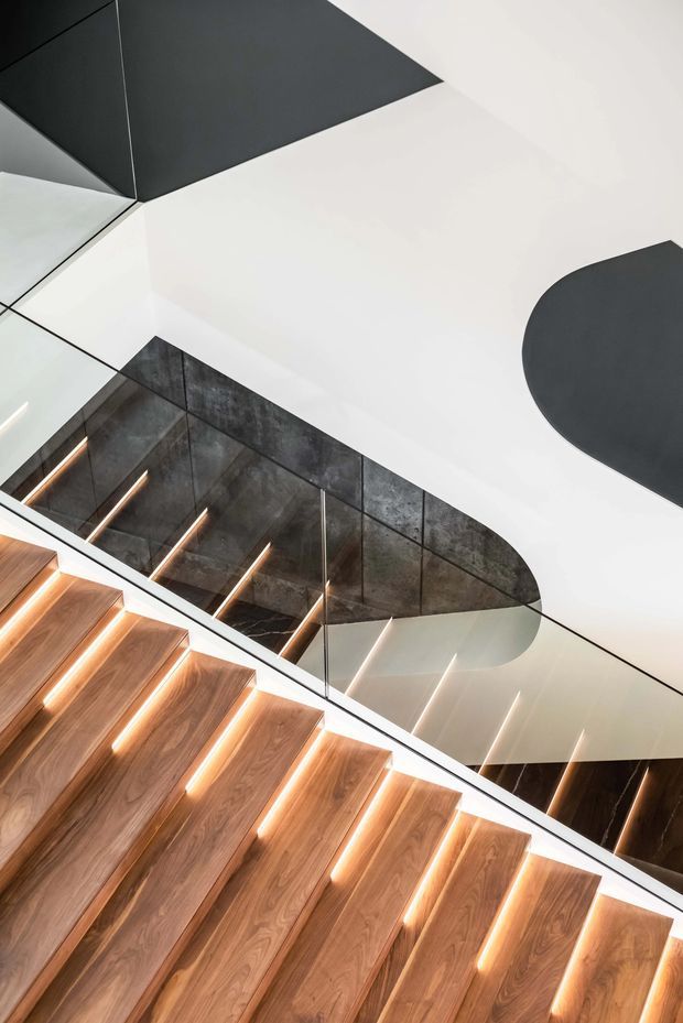 A dramatic close-up architectural view of a modern stairway, looking upward along warm timber treads with integrated linear step lighting. A clear glass balustrade and dark reflective wall panels frame the stairs, while the white ceiling features bold black geometric and curved forms. The composition is highly contemporary and sculptural, with strong contrast between the warm timber, dark metal-like surfaces, and white ceiling.