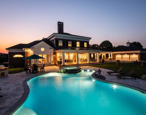A large traditional-style suburban home photographed at dusk from the pool area. The two-storey central section has dark painted siding, white trim, a prominent brick chimney, and illuminated windows, with single-storey wings extending on both sides. A curved, brightly lit swimming pool with a raised circular spa dominates the foreground, surrounded by pale paved terraces, outdoor seating, dining furniture, umbrellas, and landscaped lawn.
