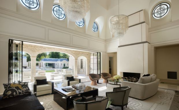 A bright, refined living room with tall paneled walls, arched openings, clerestory-style round windows, and large open steel-framed doors leading to a formal outdoor courtyard. The room features sculptural chandeliers, a fireplace and built-in cabinetry, contemporary sofas and armchairs, a glass coffee table, and a neutral cream, white, and black palette. Decorative geometric window glazing and ornate architectural detailing create a luxurious classic-contemporary atmosphere.