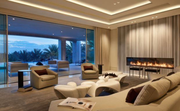Luxurious contemporary living room with a large cream sectional sofa, sculptural cream lounge chairs and coffee tables, floor-to-ceiling glazing opening to a tropical exterior, and a long linear fireplace set within vertically textured wall panelling. Layered recessed ceiling details and concealed cove lighting create a refined, warm atmosphere.
