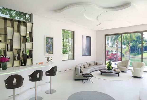 Bright, spacious contemporary open-plan living area with a white bar counter and sculptural dark bar stools, a curved sectional sofa, modern lounge chairs, and a low coffee table. Floor-to-ceiling glazing opens views to a landscaped garden. The room features a sculptural white ceiling installation, integrated display shelving, pale flooring, and minimalist artwork.