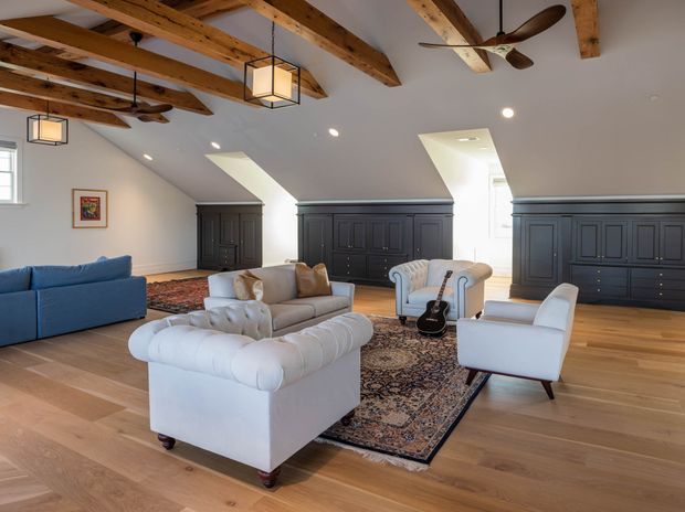 A spacious attic-style living room with exposed timber roof beams, sloped ceilings, warm timber flooring, and dark built-in panelled cabinetry along the rear wall. The room is furnished with white and blue sofas and armchairs arranged around patterned area rugs, with pendant lights, recessed lighting, skylights, and ceiling fans. The overall character blends traditional architectural detailing with refined contemporary furnishings.