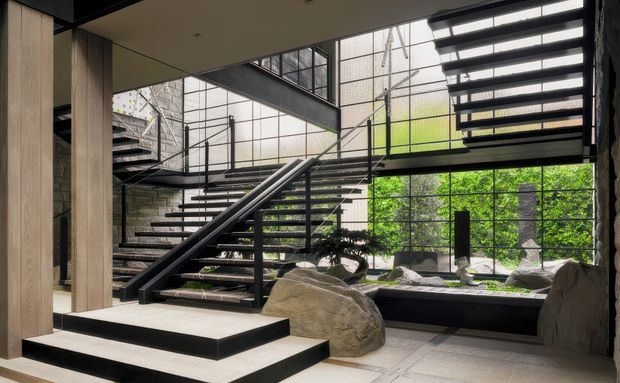 A dramatic contemporary interior centered on a sculptural open-riser staircase with black metal stringers, railings, and treads. Full-height glazed walls overlook a lush planted courtyard, while large natural boulders and a shallow water or gravel feature sit beneath the stairs. Pale timber wall cladding and light stone or concrete floor tiles contrast with the dark stair structure. The space appears tall and gallery-like, with an additional upper-level walkway visible.