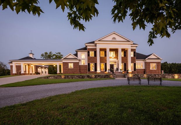 A large symmetrical brick-and-stone residence photographed at dusk, featuring a prominent classical-style portico with tall white columns and a triangular pediment, flanking wings, illuminated windows, and a separate covered porte-cochère on the left. The building sits within expansive landscaped grounds with a curved gravel driveway and mature trees framing the view.