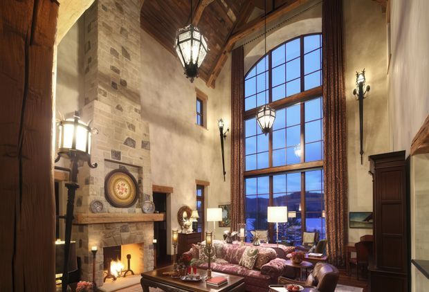 A dramatic, richly detailed living room with a large stone fireplace, exposed timber vaulted ceiling, tall timber-framed multi-pane glazing, ornate lantern and wall lighting, and traditional dark wood furnishings. The space has a warm, lodge-like character with a view through the windows to distant mountains or hills.