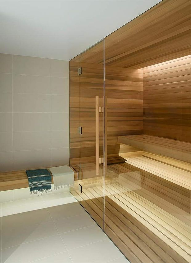 A contemporary sauna and adjacent tiled wet area. The sauna is lined with warm horizontal timber boards and includes tiered timber benches, integrated recessed lighting, and a clear frameless glass door with a timber handle. A pale tiled wall and floor sit alongside the sauna, with a small bench and folded towel visible.