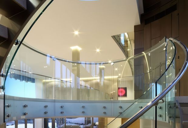 A sophisticated contemporary interior focused on a curved staircase with clear glass balustrades and a polished metal handrail. The stair overlooks a bright, open multi-level space with extensive glazing, recessed lighting, pale surfaces, and dark timber wall panelling. A partial lounge area is visible below and through the glass.