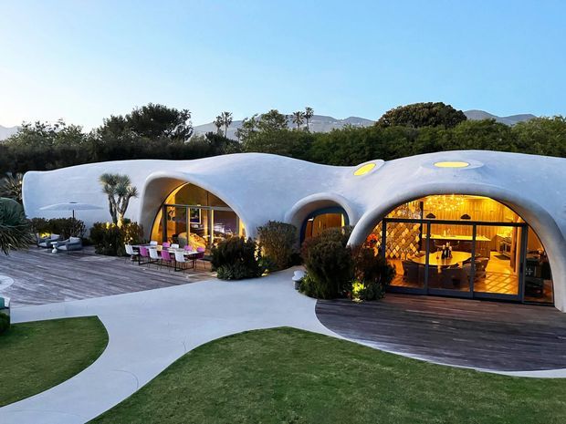 Exterior view of a low, organic sculptural building with smooth white rounded walls, arched openings, and oval skylights. Warmly lit interiors are visible through extensive glazing, with timber decking, curved concrete paths, landscaped lawns, shrubs, and palm trees in the foreground. Mountainous terrain is visible in the distance.
