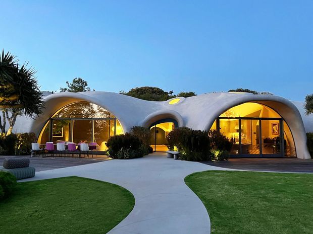 Exterior view of a sculptural, organically curved single-storey residence with a smooth white shell-like facade and rounded arched openings. Warm interior lighting glows through expansive floor-to-ceiling glazing, with outdoor dining furniture, manicured lawns, curved concrete paths, and landscaped planting in the foreground. The image is taken at dusk under a clear blue sky.