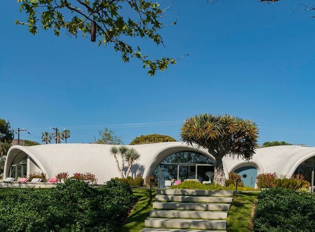 Single-storey sculptural residence with smooth white, organically curved arch forms and large glazed openings, approached by broad concrete steps through a landscaped front garden. The building has a futuristic, low-profile appearance beneath a clear blue sky, with rounded roof and facade elements defining the architecture.