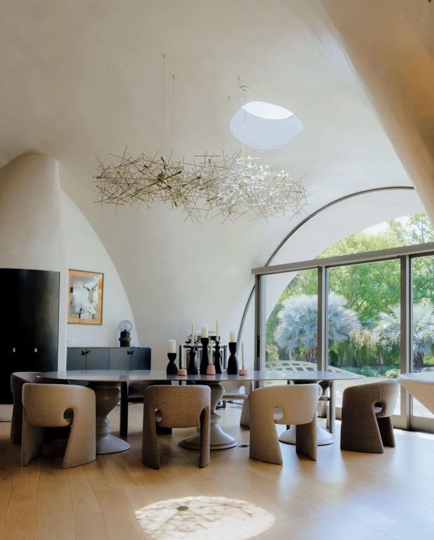 A bright, sculptural dining area with a long dark dining table surrounded by rounded upholstered chairs. The room features a dramatic curved white ceiling and arched window wall, pale timber flooring, a black media cabinet and television, restrained decorative objects, and a delicate linear pendant light. Sunlight enters through the large glazed opening and highlights the airy, refined interior.