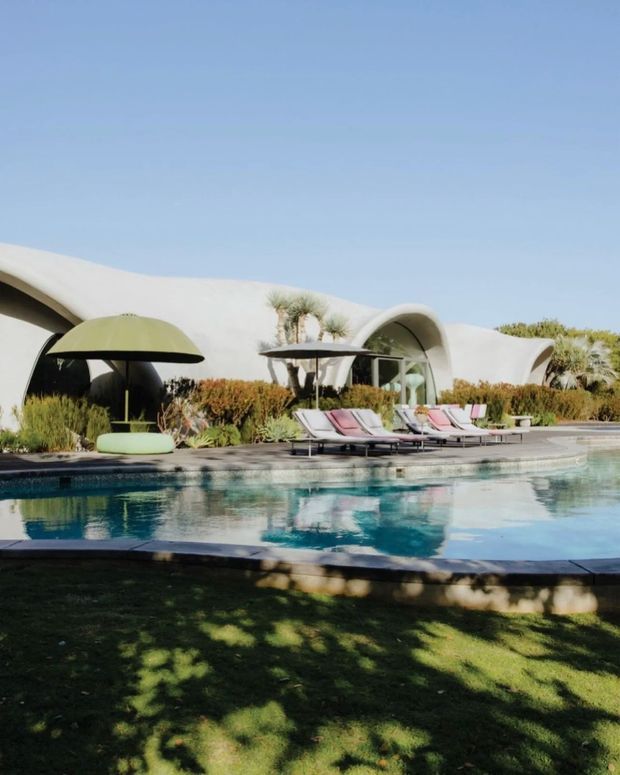 Exterior view of a sculptural low-rise residence beside a swimming pool. The building has smooth white curved walls and vaulted, shell-like roof forms, with extensive glazing and a landscaped pool terrace furnished with sun loungers and curved shade structures. A manicured lawn occupies the foreground under a clear blue sky.