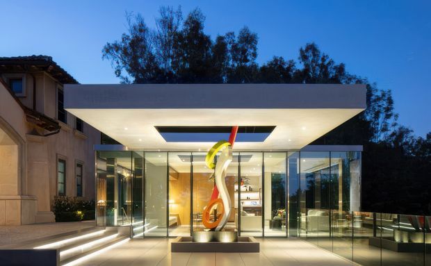 Twilight exterior view of a contemporary glass pavilion or extension attached to a more traditional rendered residence. The addition has extensive floor-to-ceiling glazing, a broad flat projecting roof, recessed ceiling lighting, a large skylight opening, and a sculptural artwork displayed in the glazed entry zone. A paved terrace and illuminated steps lead to the pavilion, with mature trees behind.
