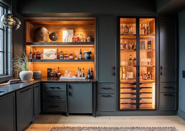 A sophisticated home bar and drinks storage area with dark charcoal cabinetry, warm timber-lined open shelving, illuminated glass-front display cabinets, and a dark stone benchtop. Bottles, glassware, decorative objects, and artwork are arranged throughout the joinery. A large window and pendant light add contrast, while timber flooring and a patterned rug soften the space.