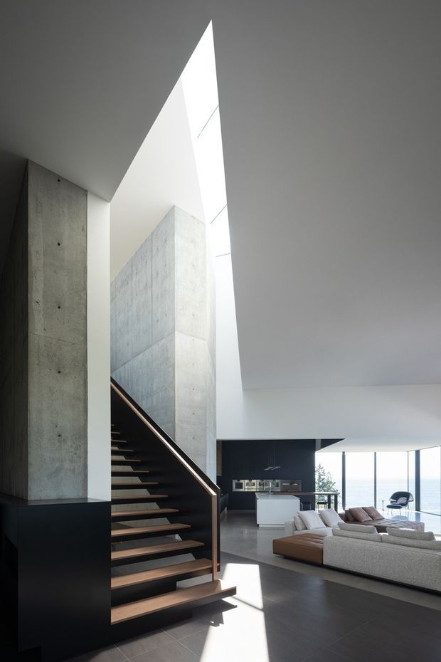 A dramatic double-height contemporary interior combining an open stairway, exposed concrete walls, dark cabinetry, and a bright open-plan living area. The stair has dark stringers and warm timber treads, rising beside a tall narrow skylight or clerestory. A large sectional sofa faces the open space, with a kitchen or bar area and floor-to-ceiling glazing visible beyond. The restrained palette and strong natural light create a refined, architectural atmosphere.